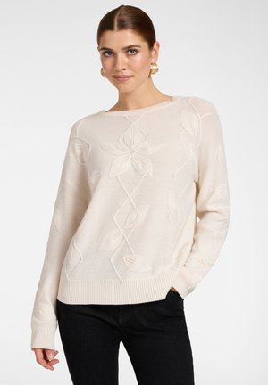 Ung kvinde med lyst beige sweater med blomsterprint og sorte bukser, poserer med den ene hånd i lommen og guldøreringe.