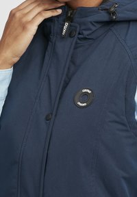 Mörkblå vadderad jacka med hög krage, dragkedja, framknappar och en OXMO-logotyp i en rund svart accent.