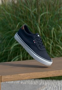 Navy canvas sneaker with white rubber sole, brown leather heel accent, and lace-up design featuring a side logo and red/white/blue stripe detail.