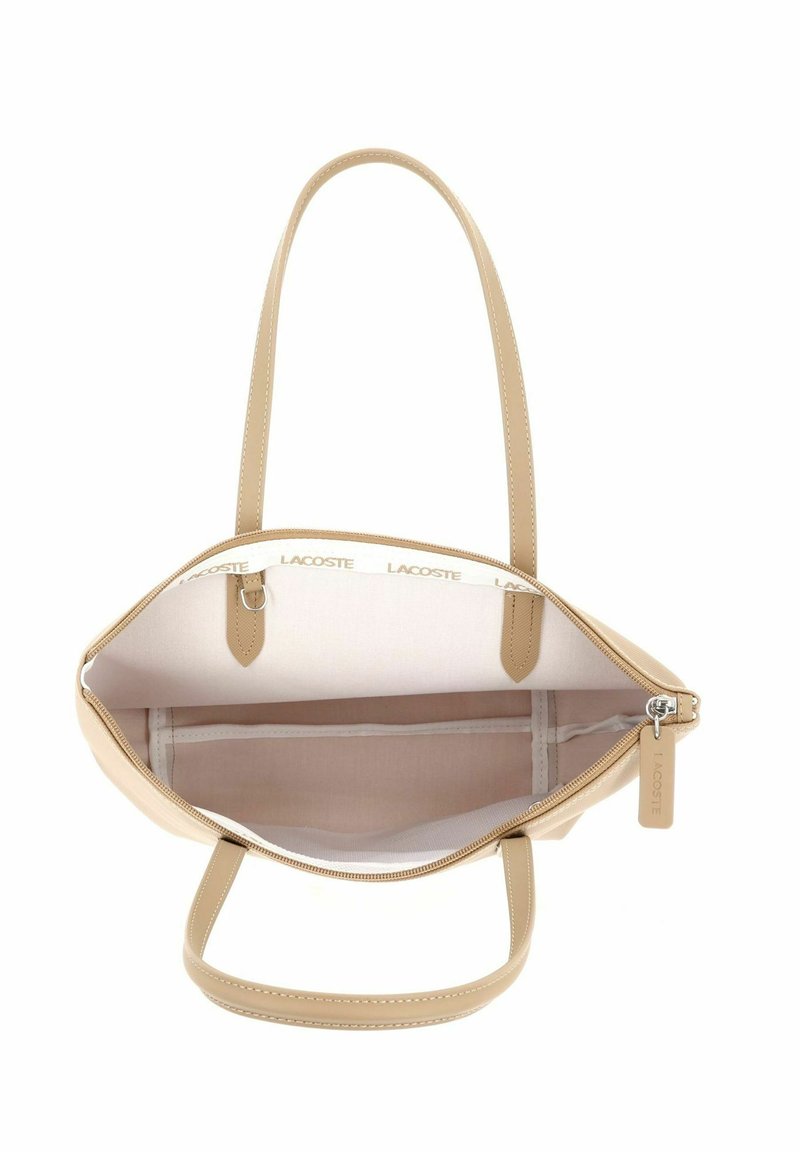 Beige handbag with two handles, open zipper showing spacious interior with side pocket and Lacoste-branded trim inside the bag.