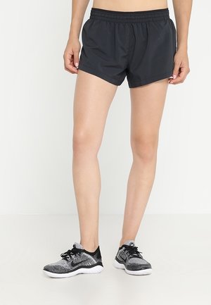 Mujer con pantalones cortos deportivos negros y zapatillas de correr negras y grises de pie sobre un fondo blanco.