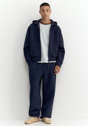 Jeune homme debout contre un mur blanc portant un sweat à capuche zippé bleu marine, un pantalon de survêtement bleu marine assorti, un t-shirt blanc et des baskets beige.