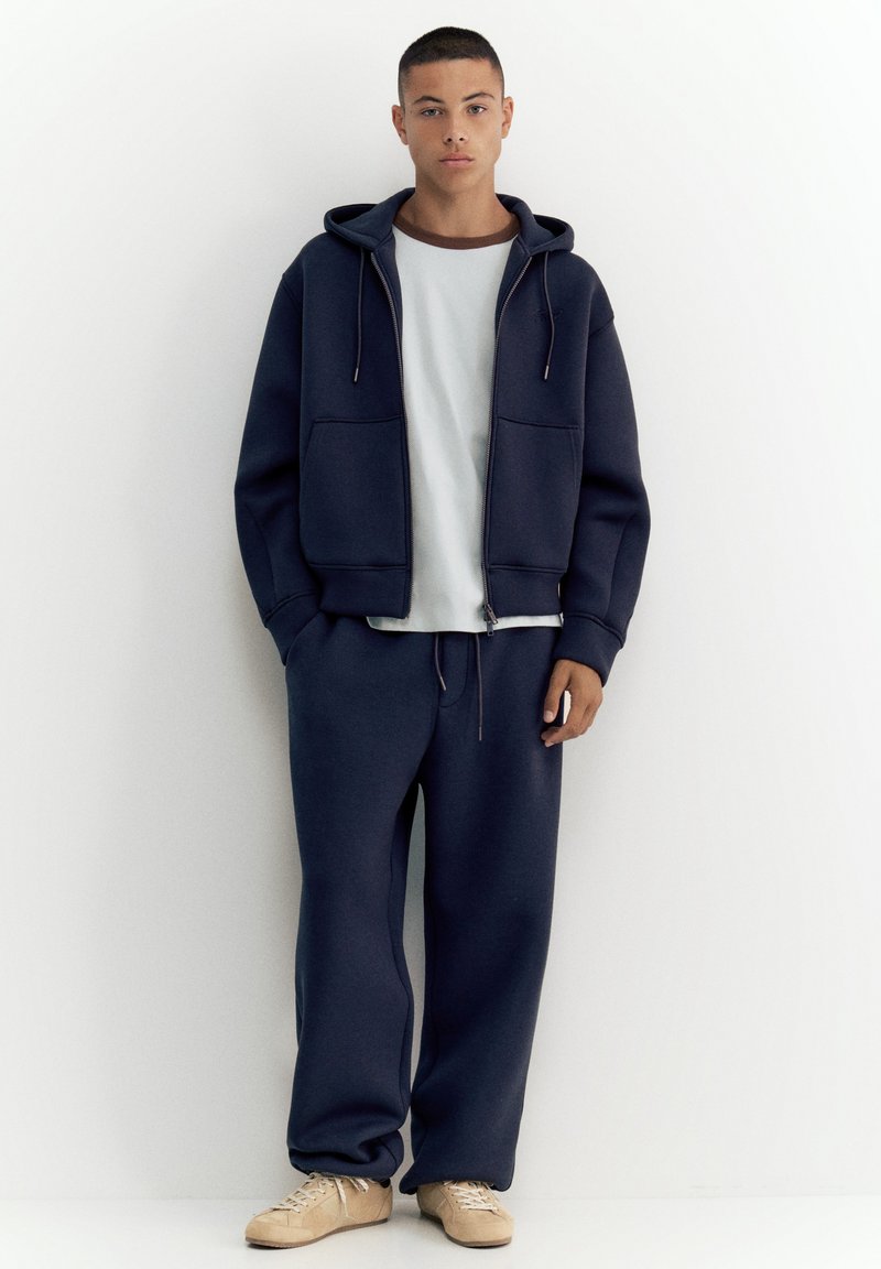 Jeune homme debout contre un mur blanc portant un sweat à capuche zippé bleu marine, un pantalon de survêtement bleu marine assorti, un t-shirt blanc et des baskets beige.