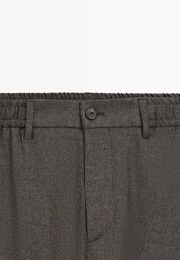 Pantaloni grigi scuri con cintura elastica, chiusura a un bottone e tasche laterali. Il tessuto ha una sottile texture e lucentezza.
