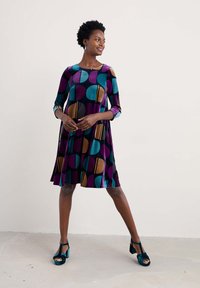Ein knie-langes Kleid mit Dreiviertelärmeln, das ein mehrfarbiges geometrisches Muster in Lila, Türkis, Gold und Schwarz aufweist. Glatter Stoff.