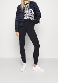 Schwarze Pufferjacke, gemusterter Pullover mit "TOMMY"-Aufdruck, schwarze Leggings und weiße Sneaker. Glatte Texturen und taillierte Passform.