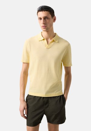 Mann trägt ein hellgelbes Polohemd mit kurzen Ärmeln, dunkle Shorts und schwarze Sonnenbrille auf dem Kopf, steht vor einfarbigem hellem Hintergrund.