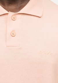 Κοντινή λήψη πουλόβερ πόλο σε χρώμα ροδάκινου με δύο κουμπιά και κεντημένο το "BOSS" στο στήθος.