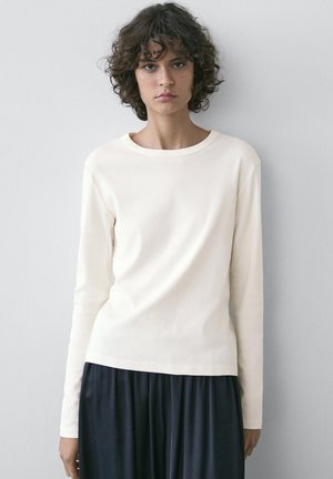 Jeune personne aux cheveux bouclés et foncés portant un haut à manches longues crème uni et un pantalon noir ample, debout contre un mur gris clair.