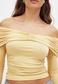 Rumena brezramenska bluzka z nabranimi detajli na prsih, dolgimi rokavi in oprijetim pasom, izdelana iz mehkega raztegljivega materiala.