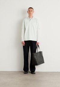 Zwarte tote bag met het reliëf "HUGO" logo, gedragen door een persoon in een lichtgroene polo en zwarte broek, staand tegen een effen muur.