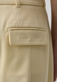 Beige trousers featuring a back pocket with a flap, embossed "SAMSØE SAMSØE" logo, smooth texture, and tailored design.