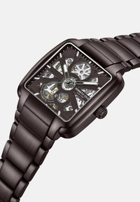 Quadratische Automatikuhr mit schwarzem Metallgehäuse und -armband. Durchsichtiger Zifferblatt zeigt die inneren Mechaniken; silberne Akzente und detaillierte Texturen.