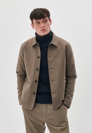 Brown cotton jacket with a collared neck, front buttons, and two side pockets, worn over a navy ribbed turtleneck sweater and corduroy pants.