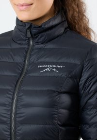 Zwart pufferjack met hoge kraag, voorzien van horizontale stiksels, een ritssluitingen aan de voorkant en een wit "SWEDEMOUNT" logo op de borst.