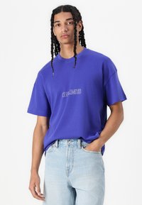 T-shirt violet oversize avec un graphique "OFF-LIMITS" en blanc. Tissu en coton, manches courtes, col rond, coupe décontractée, associé à un jean en denim bleu clair.