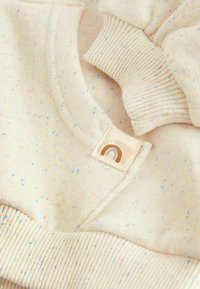 Sweatshirt beige clair avec des éclats multicolores. Il présente des poignets et un ourlet côtelés, avec une petite étiquette en tissu affichant un motif arc-en-ciel.