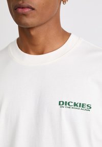 Tricou din bumbac de culoare crem, cu un text grafic verde pe pieptul stâng, cu guler rotund și mâneci scurte.