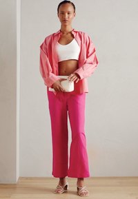Rosa oversized skjorte med knapper, hvit crop top, knallrosa vide bukser og hvit håndveske. Modell som har på seg strappy hvite sandaler.