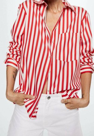 Persoon draagt een rood-wit verticaal gestreept overhemd met opgerolde mouwen en witte broek, handen in de zakken.