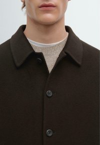 Cappotto di lana marrone scuro con colletto classico. Presenta bottoni neri e un maglione beige a trama sotto. Design minimalista.