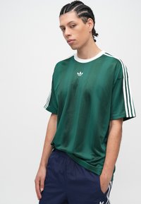 Πράσινη φανέλα με κοντά μανίκια και λευκή λαιμόκοψη, με κάθετες ρίγες και τρεις λευκές ρίγες στα μανίκια, με το λογότυπο της Adidas στο κέντρο.