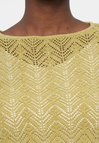 Κοντινό πλάνο ενός λαιμού και του άνω μέρους του στήθους ενός ατόμου που φοράει ένα ανοιχτό πράσινο πλεκτό ένδυμα με σχέδιο ζιγκ-ζαγκ από δαντέλα.