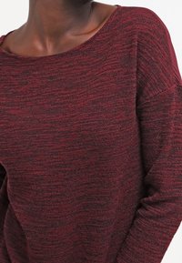 Maglione a maglia bordeaux con superficie testurizzata, scollatura ampia e maniche lunghe. Presenta lievi variazioni di colore e una vestibilità rilassata.