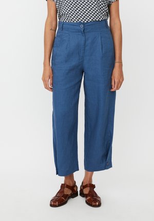 Pantaloni azzurri in lino ampi e corti con vita alta, pieghe frontali e dettagli laterali con bottoni. Abbinati a sandali marroni con cinturini.