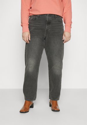 Personne portant un jean noir délavé, des bottines marron à talons et un haut rose corail à manches longues, debout sur un fond blanc.