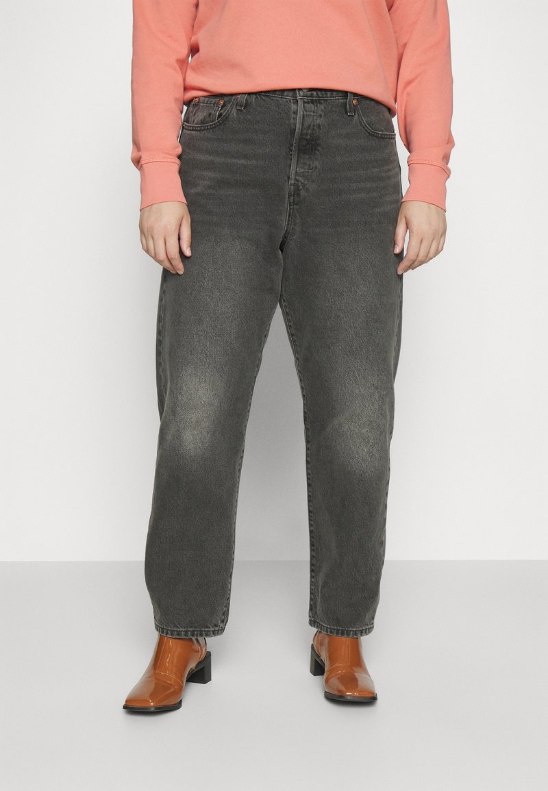 Personne portant un jean noir délavé, des bottines marron à talons et un haut rose corail à manches longues, debout sur un fond blanc.