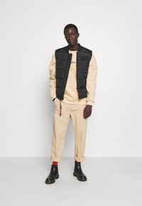 Svart pufferväst över beige sweatshirt, beige uppvikta byxor och svarta ankelstövlar med röda strumpor. Enkel design, avslappnad passform.