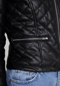 Veste en cuir noir matelassée avec un gros plan sur la poche zippée et la finition texturée. Présente des coutures diagonales et une coupe slim.