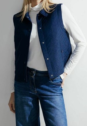 Kobieta ubrana w granatową pikowaną kamizelkę na białym, długim golfie z długim rękawem i niebieskich dżinsach, stojąca z jedną ręką w kieszeni.