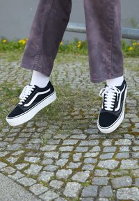 Osoba w czarno-białych sneakersach i szarych spodniach z białymi skarpetkami, stopami lekko uniesionymi nad brukowaną nawierzchnią w pobliżu metalowego słupka.