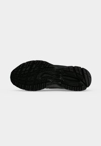 Semelle noire d’une chaussure de sport avec des motifs de bande de roulement profonds et des grips texturés sur un fond clair uni.
