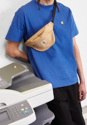 Blå t-shirt med rund hals, tanfarvet crossbody-taske med logo, sorte bukser, placeret ved siden af en grå multifunktionsprinter med kontrolpanel.