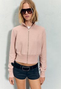 Roza zip-up skrajna bluza s strukom, nošena preko bele majice. Model jo kombinira s temnimi kratkimi hlačami in črnim pasom. Temna sončna očala.
