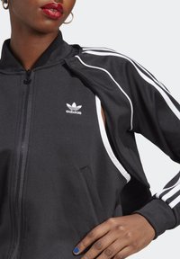 Schwarze Zip-Jacke aus einem Gewebe mit glatter Textur. Verfügt über weiße Seitenstreifen und ein gesticktes Adidas-Logo auf der Brust.