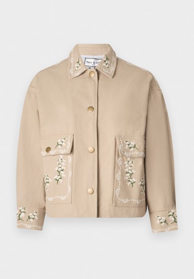 Giacca beige con colletto e ricami floreali. La parte anteriore presenta due ampie tasche, bottoni dorati e cuciture decorative sulle maniche.