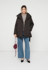 Brun polstret jakke med stor krage, knappelukking og snor. Sammen med mønstret genser og vide blå jeans. Svarte sko.