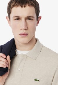 Beige bomullspolo med en grön krokodil-logotyp, som har en tre-knapplånging och texturerad tyg, bärs med en marinblå jacka över axeln.