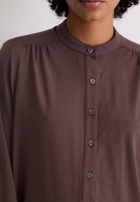 Chemise marron à manches longues avec un col rond, cinq boutons à l'avant et des plis subtils aux épaules, fabriquée en tissu texturé doux.