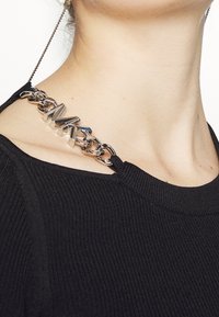 Czarna ribbed tkanina z metalowym detalem łańcucha z wypukłymi literami. Łańcuch łączy się z kołnierzem, dodając tekstury i zainteresowania.