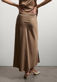 Jupe maxi en satin marron avec une taille sans couture, une silhouette fluide et un ourlet légèrement asymétrique. Texture brillante et drapé doux.