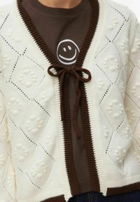 Cardigan crème avec motif en losange, boules texturées, et bordure marron. Noué à la taille avec un ruban marron assorti, porté sur une chemise sombre.