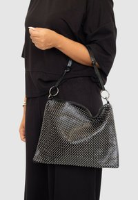 Bolso de mano negro con textura de tachuelas, que presenta una parte superior curva y dos correas con acentos de hardware circulares.