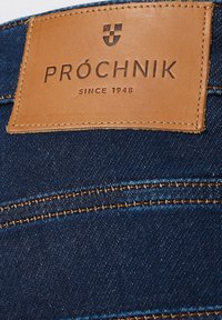 Jeansowa tkanina w ciemnoniebieskim kolorze z widocznymi szwami; ma brązową skórzaną metkę wytłoczoną z napisem "PRÓCHNIK" i "SINCE 1948."