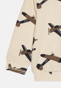 Kremfarget stoff med et helmønster av brune og svarte fly. Teksturen er ribbet, med tettsittende mansjetter og en avslappet fasong.