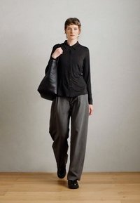 Musthave musta nööpidega pikkade varrukatega särk, hall triibuline laiad püksid, mustad kingad ja suur must kott, neutraalses taustas.